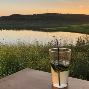 Coucher de soleil sur un lac calme depuis l'intérieur d'un van aménagé, avec un verre de cocktail au premier plan
