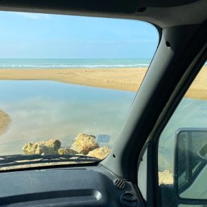 Paysage ensoleillé avec plage de sable et mer turquoise, vue depuis l'intérieur d'un van à travers le pare-brise